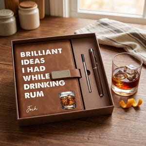 Könnte beinhalten: Ein braunes Lederjournal mit dem Text "BRILLIANT IDEAS I HAD WHILE DRINKING RUM" und einer kleinen Abbildung eines Getränks. Das Journal befindet sich in einer Geschenkbox mit einem Stift und einem Glas Rum mit Eis.