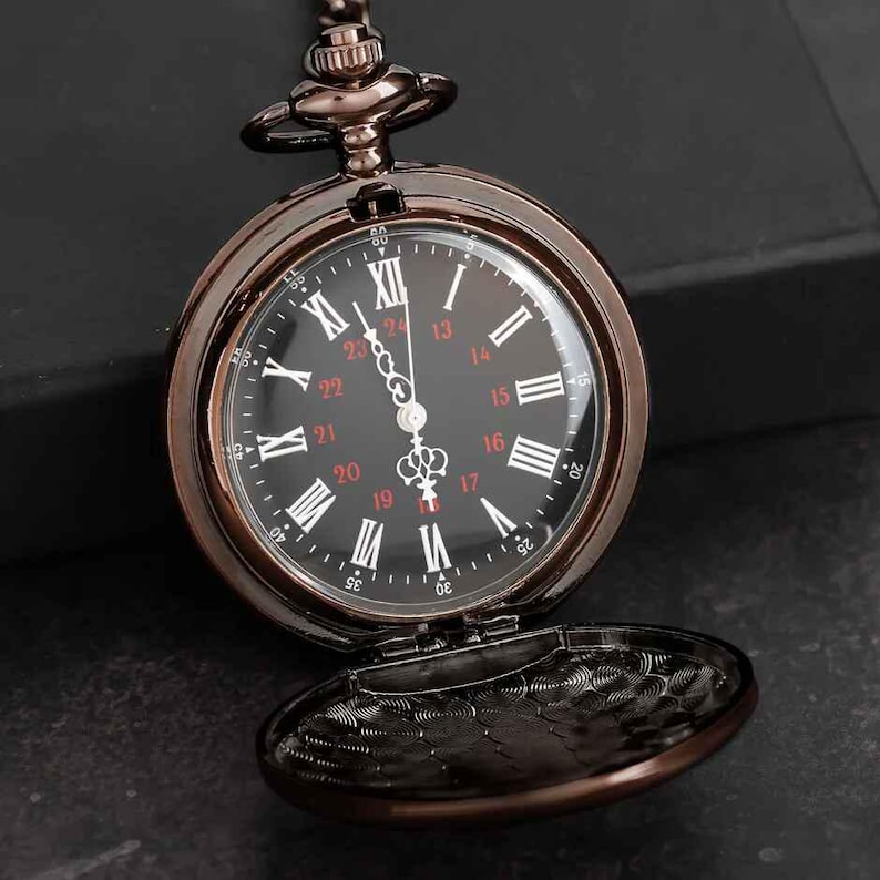 Puede incluir: Un reloj de bolsillo de color bronce con esfera negra y n&uacute;meros romanos blancos. El reloj tiene una tapa con bisagras y las manecillas indican la hora. El reloj est&aacute; sobre una superficie oscura.