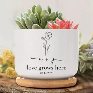 Peut inclure: Pot de fleurs en céramique blanche avec un motif floral et le texte « love grows here » avec la date du 14.02.2023. Le pot contient des succulentes vertes et rouges et repose sur un dessous de plat rond en bambou.