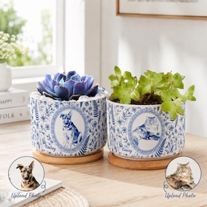 Peut inclure: Deux pots de fleurs cylindriques en céramique avec un motif floral bleu et blanc. Chaque pot présente un portrait ovale central d'un animal, avec les noms Bailey et Daisy inscrits dessous. Ils reposent sur une base en bois et contiennent une plante.