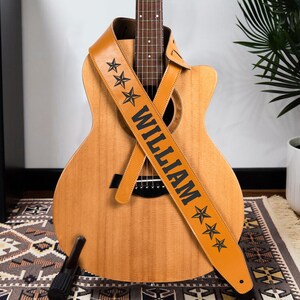 May include: A light brown guitar strap draped over an acoustic guitar. The strap features the name WILLIAM in bold black capital letters, flanked by three black star icons on each side. The guitar has a natural wood finish and a dark bridge.