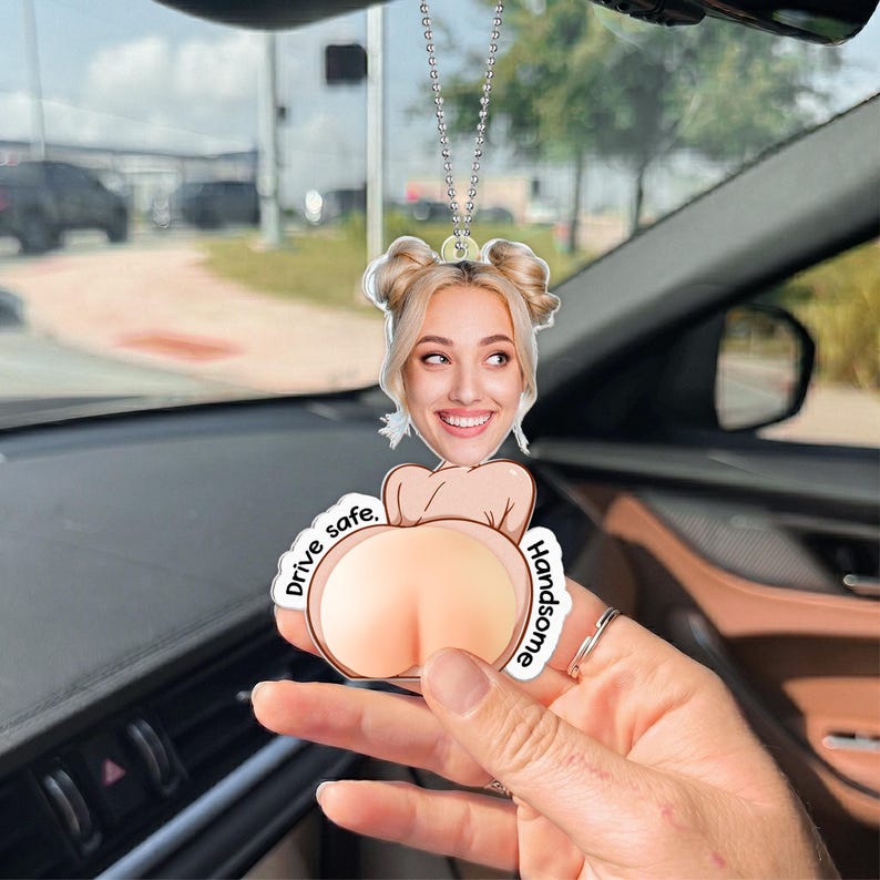 May include: A custom car air freshener hanging from a silver ball chain, featuring a printed portrait of a smiling person with blonde hair in two buns, paired with a cartoon illustration of buttocks and the text Drive safe, Handsome.