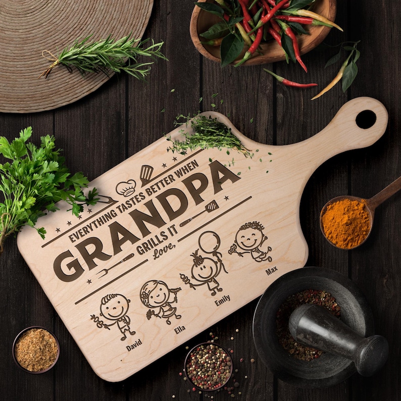 Może przedstawiać: Drewniana deska do krojenia z uchwytem, ​​z wygrawerowanymi słowami "GRANDPA GRILLS IT" i imionami wnuk&oacute;w. Deska otoczona jest ziołami, przyprawami oraz moździerzem i tłuczkiem, sugerując motyw grillowania.