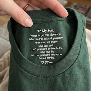 May include: A dark green t-shirt featuring white printed text on the inside collar area that reads: To My Son, Never forget that I love you. When life tries to knock you down remember I will always have your back. I can't promise to be here for the rest of your life, but I can promise to love you for the rest of mine. Mom.