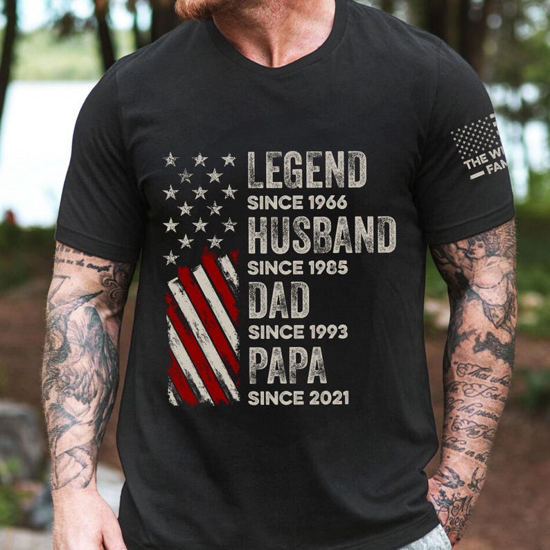 May include: Black t-shirt with a distressed American flag design. The shirt features the words "LEGEND SINCE 1966, HUSBAND SINCE 1985, DAD SINCE 1993, PAPA SINCE 2021" in white lettering. The text "THE W FAM" is on the right sleeve.