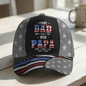 May include: A black baseball cap with a denim-style pattern and white stars. The cap features the text "FIRST DAD NOW PAPA" in red and blue, along with names. The brim displays a stylized American flag.