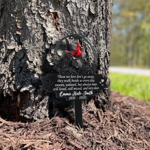 May include: A black metal memorial stake with a red cardinal perched on a branch. The stake has a circular design with floral accents and a banner with the inscription "Those we love don't go away..." and the name "Emma Hale Smith 1936-2026."