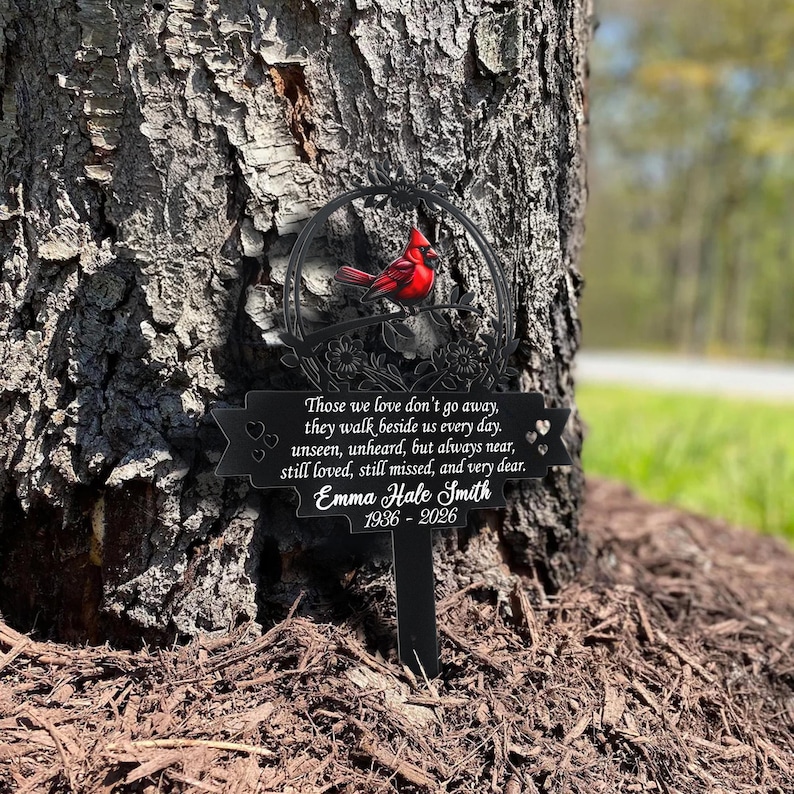 May include: A black metal memorial stake with a red cardinal perched on a branch. The stake has a circular design with floral accents and a banner with the inscription "Those we love don't go away..." and the name "Emma Hale Smith 1936-2026."
