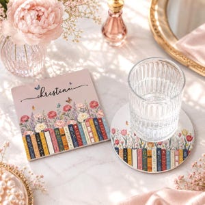 Peut inclure: Un dessous de verre carré et un rond présentant une illustration colorée d'une rangée de livres surmontée de fleurs roses et blanches et de papillons. Le dessous de verre carré affiche le prénom Christine en écriture cursive noire.
