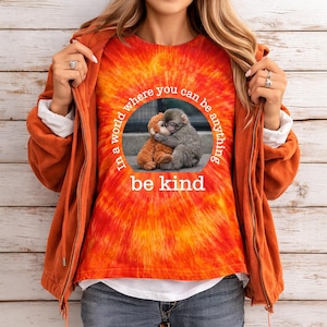 May include: An orange and yellow tie-dye t-shirt featuring the text "In a world where you can be anything, be kind" around an image of two embracing monkeys. The shirt is part of a casual outfit.