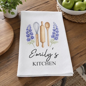 May include: A white waffle-weave kitchen towel featuring a printed illustration of a whisk, a wooden rolling pin, and a wooden spoon flanked by purple delphinium flowers. The text "Emily's KITCHEN" is printed in black below the kitchen tools.