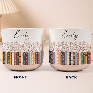 May include: Two ceramic mugs featuring the name Emily in black script above a colorful illustration of a row of books and pink wildflowers. The mugs have a speckled white upper section and a textured, tan-colored base. The text FRONT and BACK appears.
