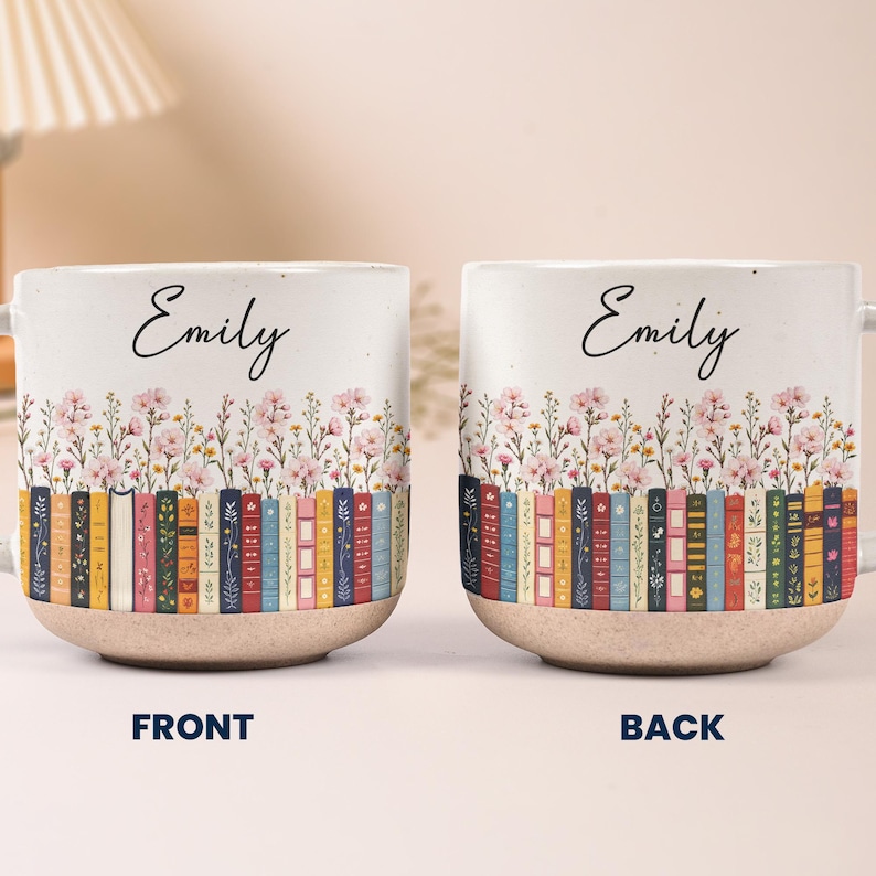 May include: Two ceramic mugs featuring the name Emily in black script above a colorful illustration of a row of books and pink wildflowers. The mugs have a speckled white upper section and a textured, tan-colored base. The text FRONT and BACK appears.