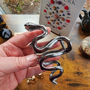 May include: A silver-colored snake-shaped bracelet with a coiled design. The bracelet features a detailed head and tail, and a spring mechanism for opening and closing. The bracelet is held in a hand, with a book and other items in the background.