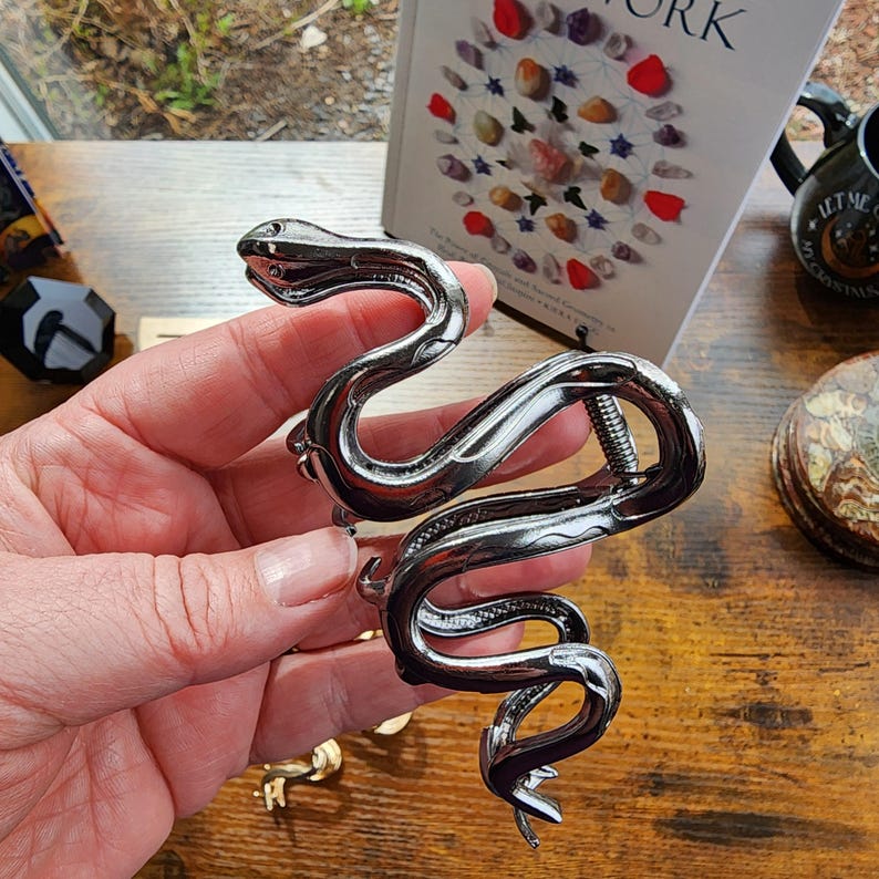 May include: A silver-colored snake-shaped bracelet with a coiled design. The bracelet features a detailed head and tail, and a spring mechanism for opening and closing. The bracelet is held in a hand, with a book and other items in the background.