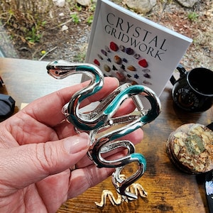May include: Silver-toned snake bracelet with teal enamel accents. The serpent design features a coiled body and a detailed head. A book titled "Crystal Gridwork" is visible in the background.
