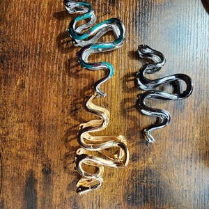 May include: Three snake-shaped hair clips in silver, gold, and black. The silver clip has teal accents, while the gold clip is a solid color. The black clip is a solid color. All three clips are on a wooden surface.