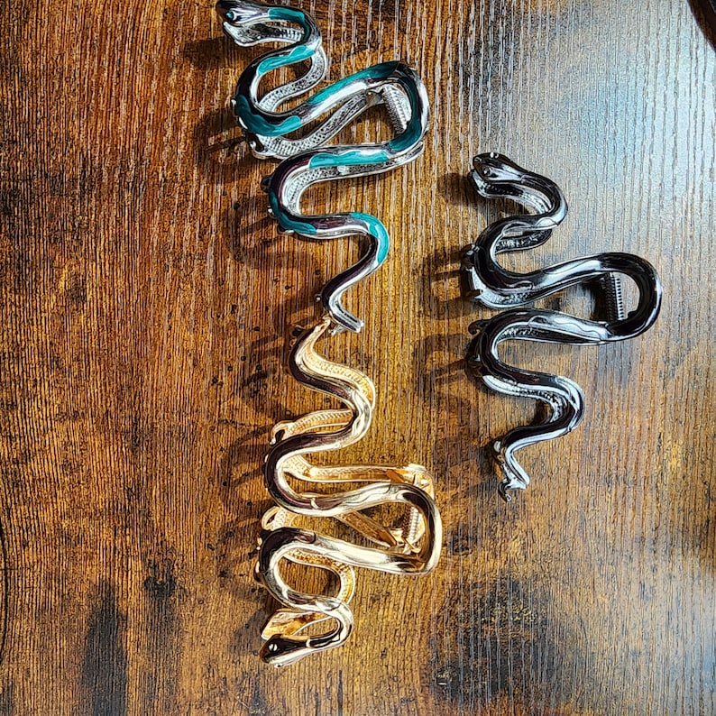 May include: Three snake-shaped hair clips in silver, gold, and black. The silver clip has teal accents, while the gold clip is a solid color. The black clip is a solid color. All three clips are on a wooden surface.