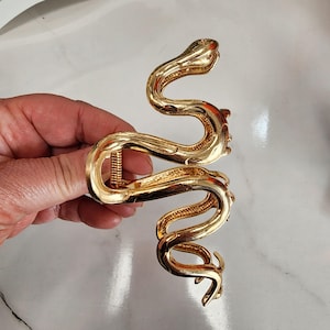 May include: A gold-colored hair claw clip in the shape of a snake. The clip is made of metal and has a detailed, textured design. The snake's body curves in an S-shape, with the head and tail forming the clip's ends. The clip is held by a hand.
