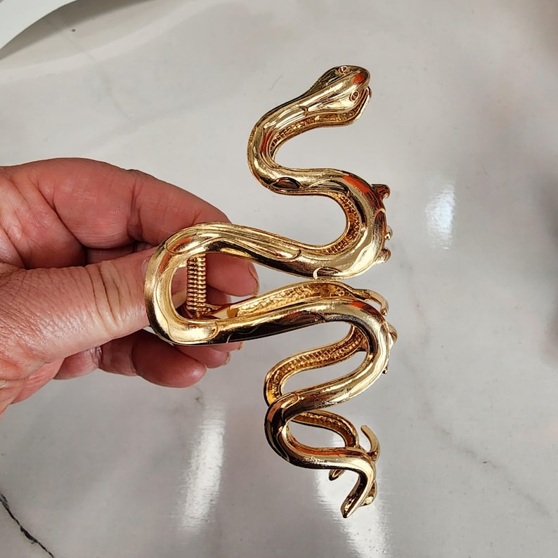 May include: A gold-colored hair claw clip in the shape of a snake. The clip is made of metal and has a detailed, textured design. The snake's body curves in an S-shape, with the head and tail forming the clip's ends. The clip is held by a hand.