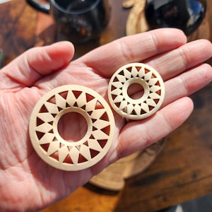 以下が含まれることがあります： 星形の幾何学模様が施された木製の円形イヤリングが2つ。イヤリングは中央に円形の穴が彫られています。イヤリングは手のひらに置かれており、左側に大きいイヤリング、右側に小さいイヤリングが置かれています。