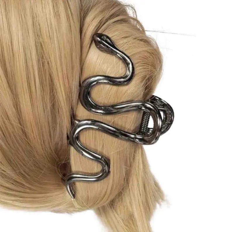 May include: A silver-colored hair clip in the shape of a snake, wrapped around a blonde hair bun. The snake's head is at the top, with the body curving down and around the hair. The clip is made of metal and has a detailed design.