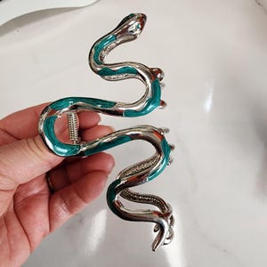 May include: A silver-toned hair clip in the shape of a snake. The snake's body is accented with teal enamel and small crystal accents. The clip is designed to hold hair in place.