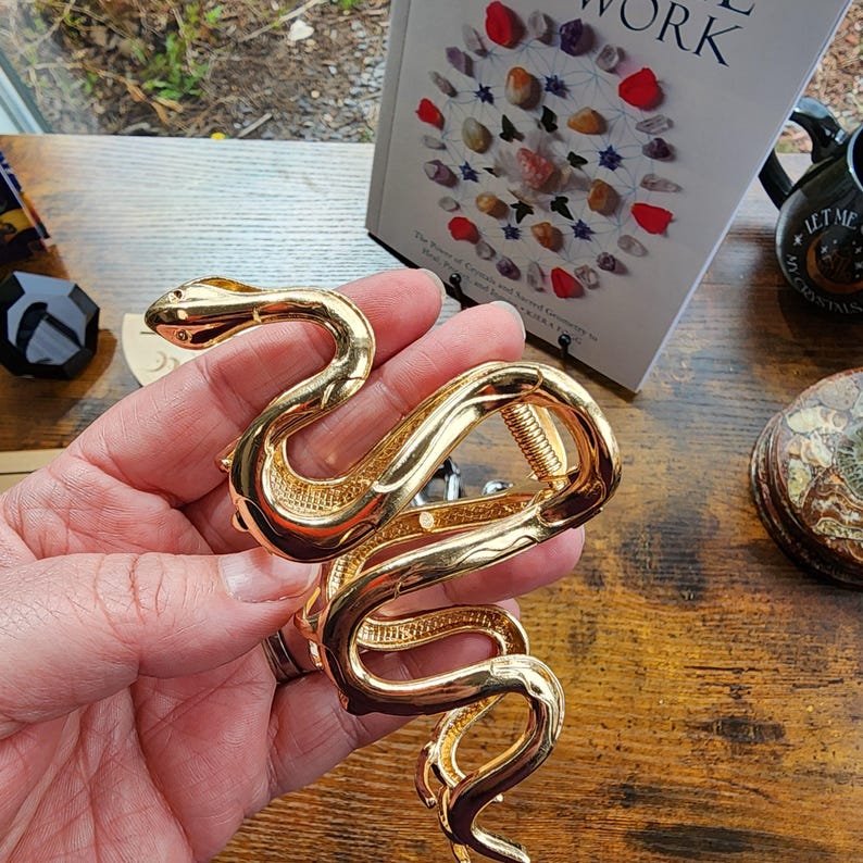 May include: A gold-colored snake bracelet with intricate detailing. The bracelet is held in a hand, showcasing its coiled design and the textured scales. The snake's head is visible, adding to the realistic appearance. The bracelet is a statement piece.