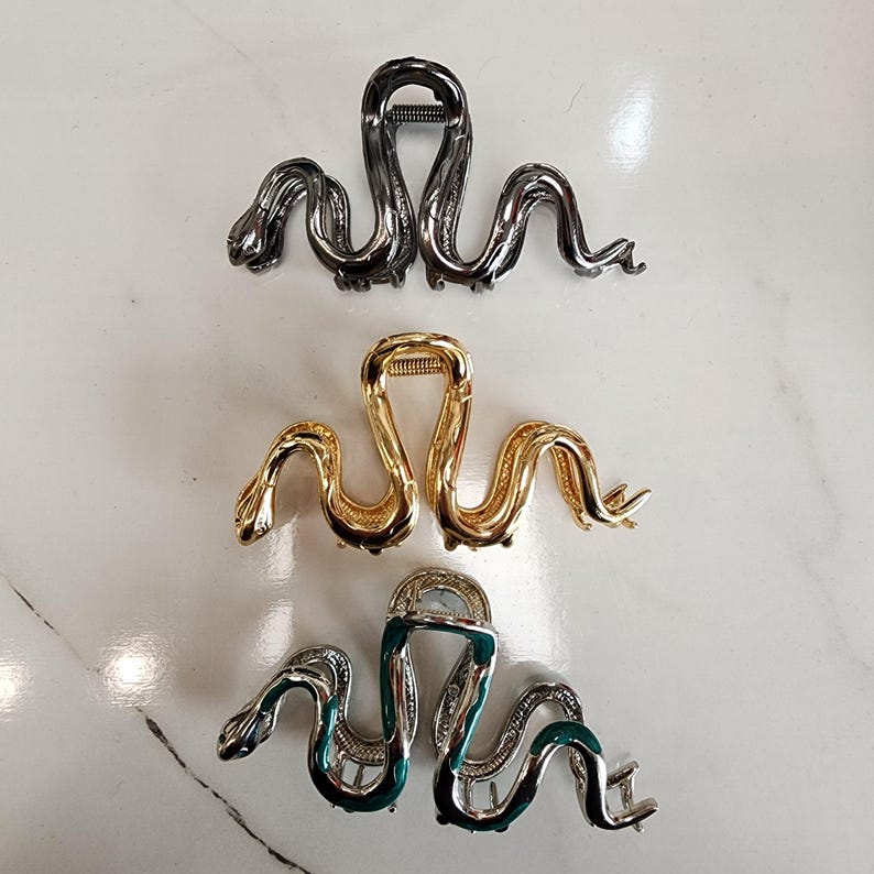May include: Three metal hair clips in a snake design. The top clip is gunmetal, the middle is gold, and the bottom clip is silver with green enamel accents. The clips are arranged on a white marble surface.