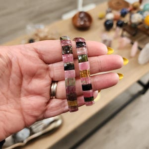 May include: Two bracelets made of multi-colored square beads. The beads are pink, green, black, and gold. The bracelets are stacked on top of each other.