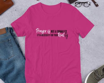 May include: A magenta t-shirt with the text "Prayer is not a formality. It's a necessity for your Reality" in white and black script. The shirt is displayed on a flat surface with jeans, a watch, and other accessories.
