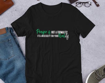 Puede incluir: Camiseta negra con texto blanco que dice "La oración no es una formalidad. Es una necesidad para tu realidad." El texto "Oración" y "realidad" están en verde.