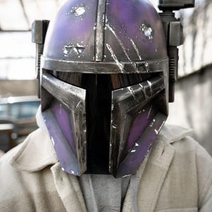 Peut inclure: Casque Mandalorien violet et argent&eacute; avec des d&eacute;tails us&eacute;s. Le casque pr&eacute;sente une visi&egrave;re en forme de T et un accessoire rectangulaire sur le c&ocirc;t&eacute; droit. Il est port&eacute; sur une veste de couleur claire et un sweat &agrave; capuche gris.