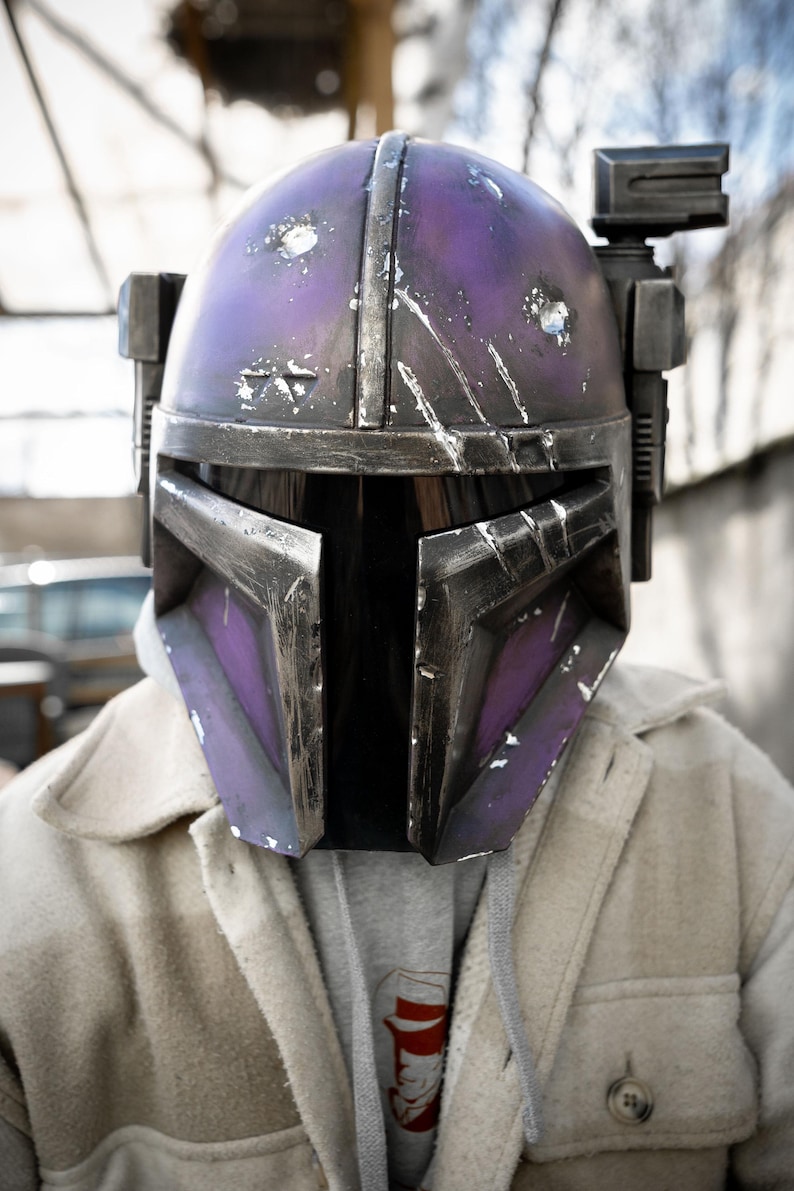 Peut inclure: Casque Mandalorien violet et argent&eacute; avec des d&eacute;tails us&eacute;s. Le casque pr&eacute;sente une visi&egrave;re en forme de T et un accessoire rectangulaire sur le c&ocirc;t&eacute; droit. Il est port&eacute; sur une veste de couleur claire et un sweat &agrave; capuche gris.