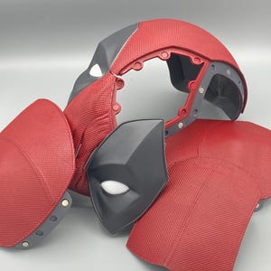 Puede incluir: Un casco de disfraz de Deadpool rojo y negro con una superficie texturizada. El casco est&aacute; en piezas e incluye una m&aacute;scara para los ojos separada.
