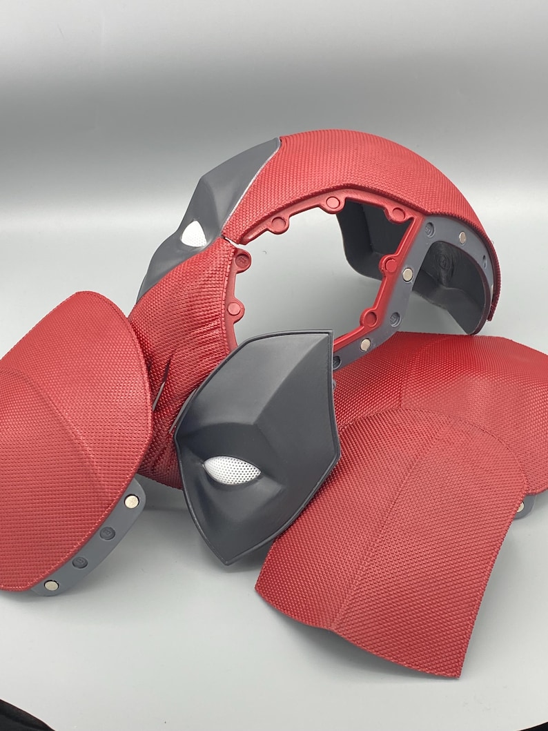 Puede incluir: Un casco de disfraz de Deadpool rojo y negro con una superficie texturizada. El casco est&aacute; en piezas e incluye una m&aacute;scara para los ojos separada.