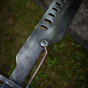 May include: A metallic blade with a weathered, dark gray finish featuring a row of rectangular cutouts along the spine. A copper-colored rod connects the blade to the handle assembly, secured by a circular silver bolt on the side of the metal.