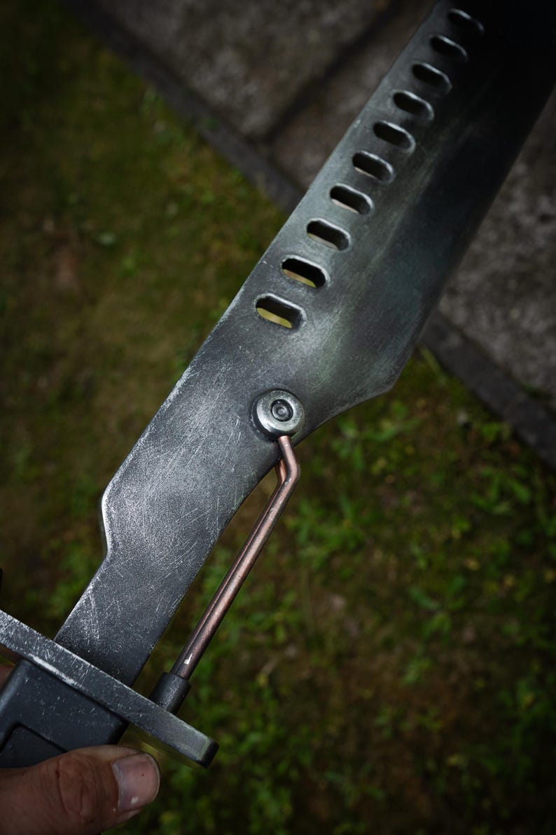 May include: A metallic blade with a weathered, dark gray finish featuring a row of rectangular cutouts along the spine. A copper-colored rod connects the blade to the handle assembly, secured by a circular silver bolt on the side of the metal.