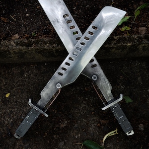 May include: Two crossed machete-style blades with a weathered metallic finish. Each blade features a series of rectangular cutouts along the spine, a dark grey handle with a rectangular grip pattern, and a small copper-colored wire detail near the hilt.