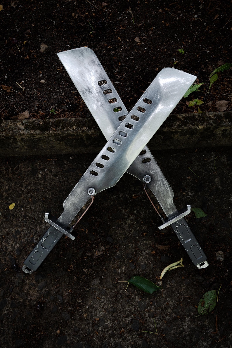 May include: Two crossed machete-style blades with a weathered metallic finish. Each blade features a series of rectangular cutouts along the spine, a dark grey handle with a rectangular grip pattern, and a small copper-colored wire detail near the hilt.