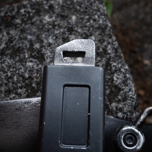 May include: A black rectangular belt buckle component with a matte finish and a metallic silver top attachment featuring a rectangular slot. The surface shows minor scuffs and wear, with a circular silver rivet visible on the lower right side.