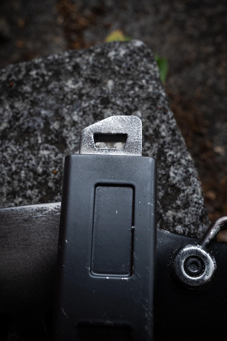 May include: A black rectangular belt buckle component with a matte finish and a metallic silver top attachment featuring a rectangular slot. The surface shows minor scuffs and wear, with a circular silver rivet visible on the lower right side.