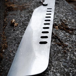 May include: A metallic blade featuring a series of ten oval cutouts along the spine. The blade has a flat, straight edge and a rounded tip, with a mechanical hinge mechanism connecting it to a dark handle visible in the background.