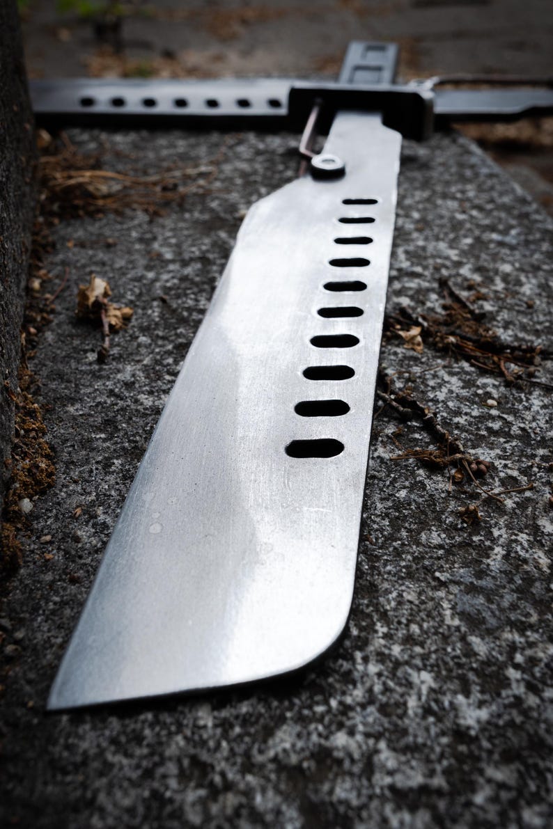 May include: A metallic blade featuring a series of ten oval cutouts along the spine. The blade has a flat, straight edge and a rounded tip, with a mechanical hinge mechanism connecting it to a dark handle visible in the background.