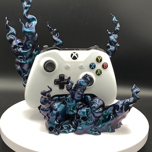 Könnte beinhalten: Ein weißer Xbox-Controller liegt auf einem Sockel mit schillernden blau-violetten Schädel- und Flammenakzenten. Der Controller hat farbige Tasten und ein schwarzes Steuerkreuz.