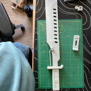 May include: A light gray 3D-printed sword prop measuring approximately 16.5 inches in length, featuring a series of rectangular cutouts along the blade, a mechanical hinge mechanism, and a rectangular handle, resting on a green cutting mat.