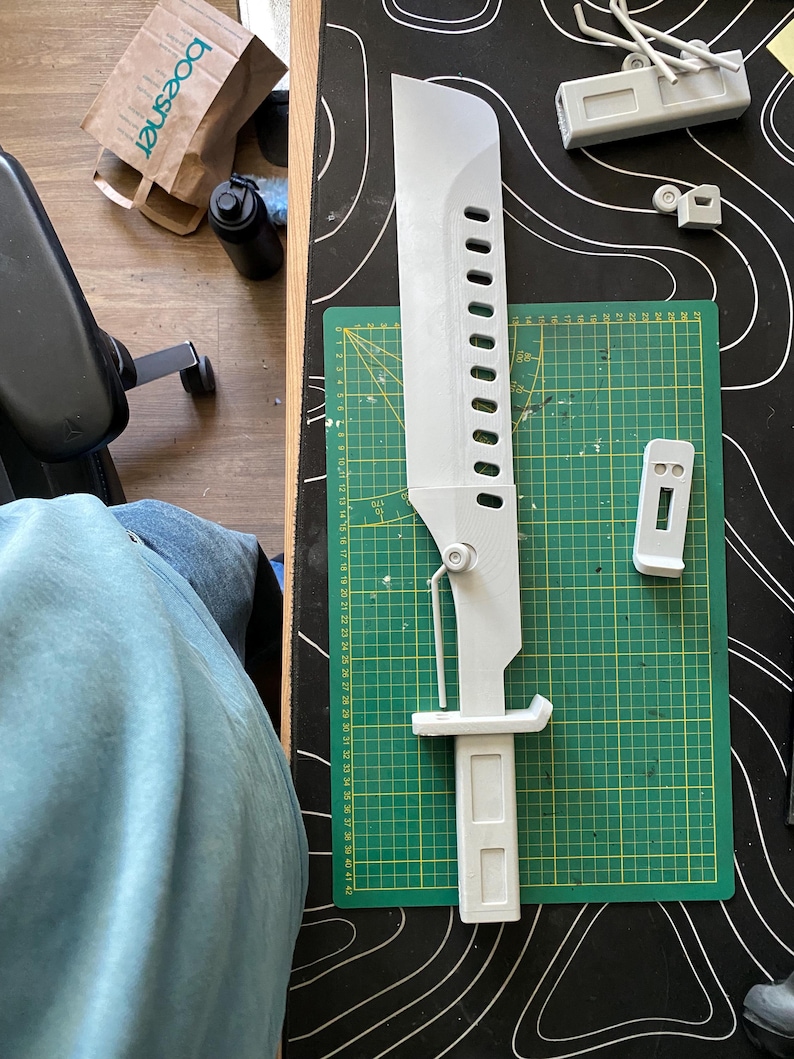 May include: A light gray 3D-printed sword prop measuring approximately 16.5 inches in length, featuring a series of rectangular cutouts along the blade, a mechanical hinge mechanism, and a rectangular handle, resting on a green cutting mat.