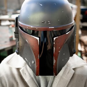 Peut inclure: Un casque Mandalorien noir et rouge &agrave; l'aspect vieilli. Le casque a une visi&egrave;re sombre et est port&eacute; sur une veste de couleur claire. Le design du casque comprend un dessus arrondi et des &eacute;l&eacute;ments angulaires.