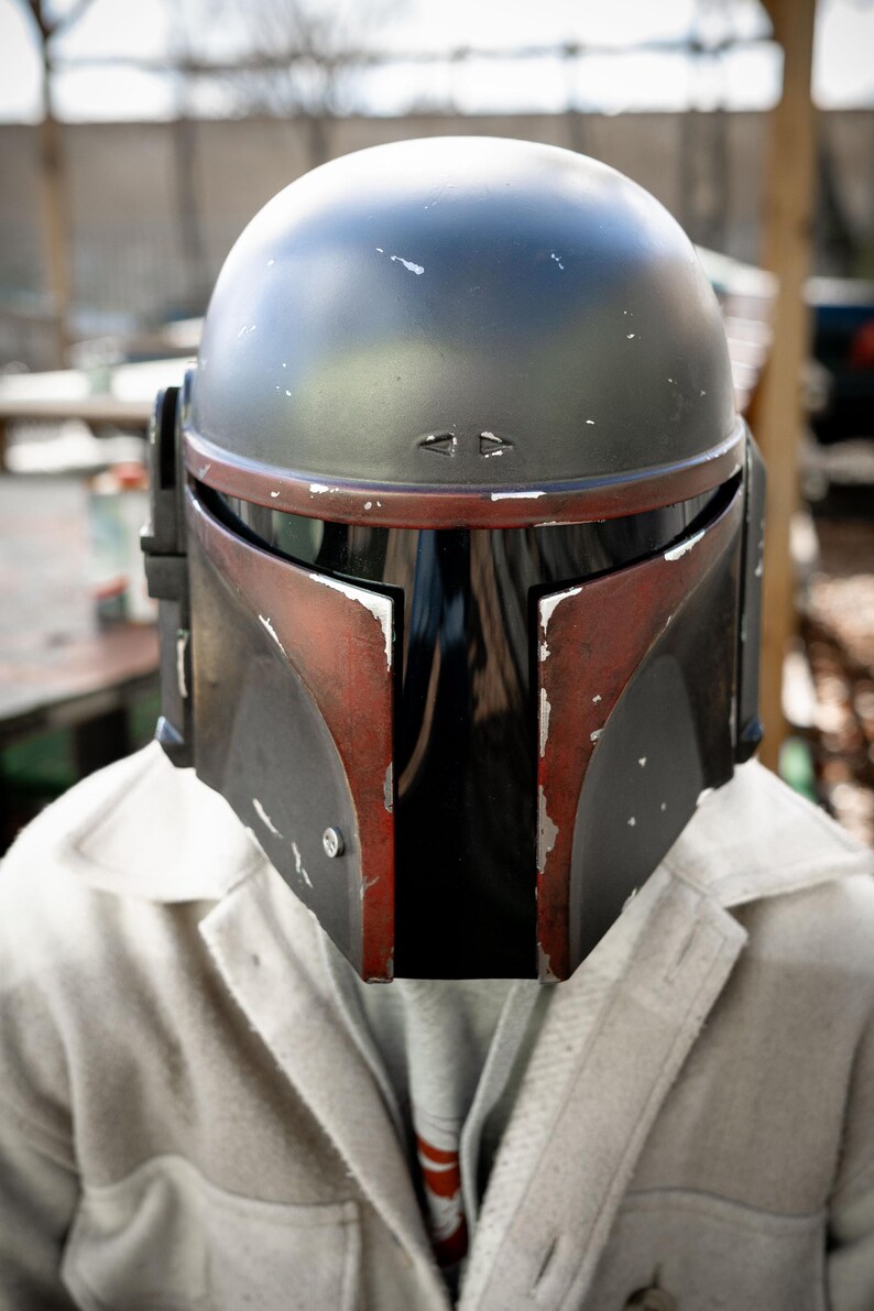 Peut inclure: Un casque Mandalorien noir et rouge &agrave; l'aspect vieilli. Le casque a une visi&egrave;re sombre et est port&eacute; sur une veste de couleur claire. Le design du casque comprend un dessus arrondi et des &eacute;l&eacute;ments angulaires.