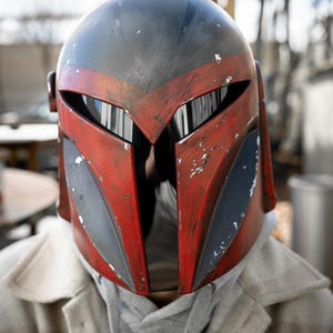 Peut inclure: Casque Mandalorien rouge et noir avec un aspect vieilli. Le casque pr&eacute;sente une visi&egrave;re en forme de T et est port&eacute; sur un sweat &agrave; capuche gris et un manteau clair. Le design du casque comprend des accents gris et noirs.